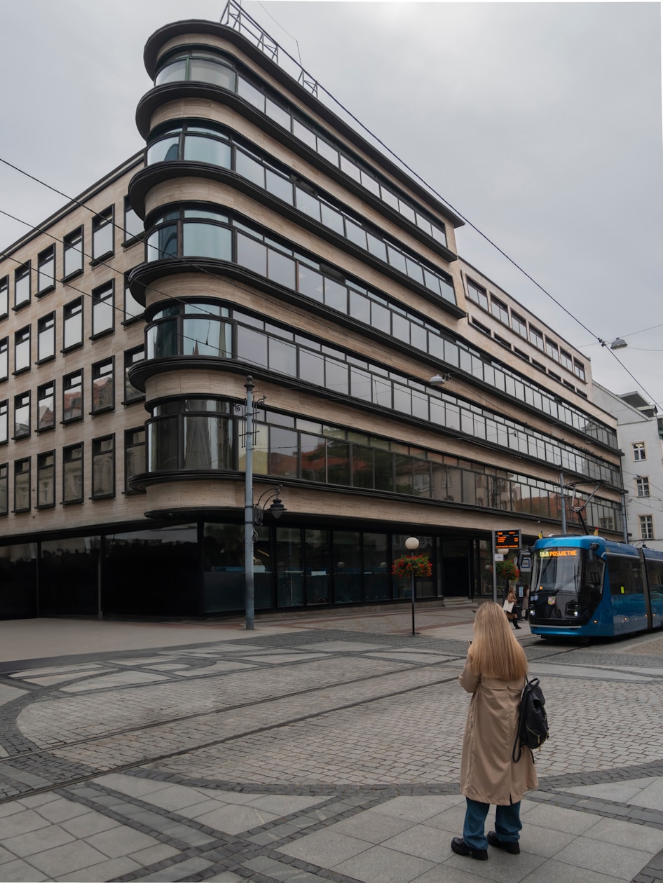 An office building with a curved corner plays backdrop to a passing blue bus and a blonde woman crossing a cobbled city street